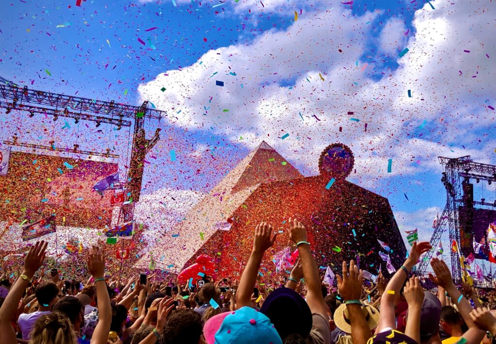 Crowd of people infront of a music festival stage, outside during the day. Confetti falling, the audiences arms are in the air. 